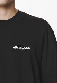 Black cotton t-shirt featuring a round neckline and short sleeves; includes a white "PEGADOR" logo on the left chest area.