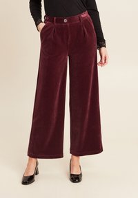 Pantaloni in velluto a coste bordeaux a gamba larga con righe verticali, caratterizzati da pieghe, chiusura con bottone e tasche laterali. Abbinati a tacchi in vernice nera.