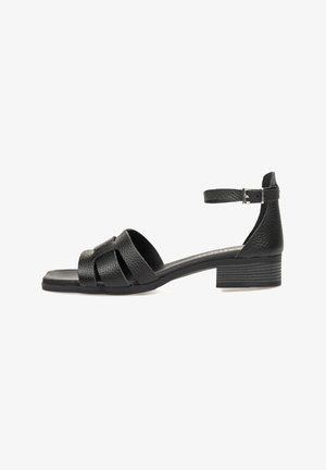 Sandalia de cuero negro con tacón bajo y cuadrado, que presenta tiras cruzadas y una correa ajustable en el tobillo. Textura suave con un acabado mate.