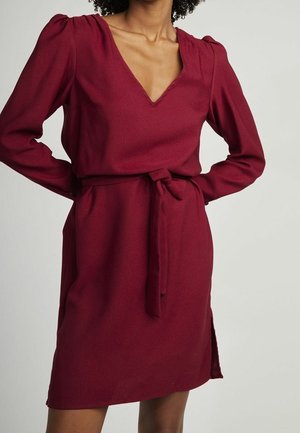 Bourgognefarvet kjole med lange ærmer og V-hals. Har en bælte ved taljen og en sideslids, lavet af tekstureret stof.