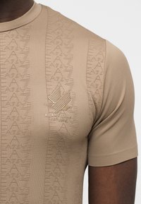 Κοντινό πλάνο μπεζ κοντομάνικου πουκάμισου με επαναλαμβανόμενο ανάγλυφο μοτίβο EA7 και κεντημένο λογότυπο Milano Italia στο στήθος.