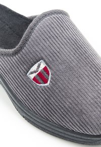 Pantofole in corduroy grigio con suola in gomma nera, caratterizzate da un logo ricamato a scudo in rosso e bianco. Texture morbida e design senza lacci.