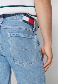 Vaqueros de mezclilla azul claro con un bolsillo trasero bordado y una etiqueta de marca visible en rojo, blanco y azul en la cintura.