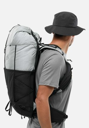 Homme portant un t-shirt gris, un chapeau large noir et un grand sac à dos de randonnée gris et noir, vu de dos sur un fond blanc.