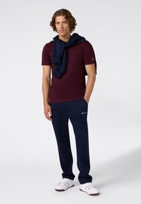 Jonge man die met zijn handen in de zakken van zijn marineblauwe sweatpants staat, gekleed in een bordeauxrood T-shirt en een marineblauwe sweater die over zijn schouders hangt, met witte sneakers.