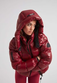 Giacca a vento rossa e lucida con un ampio cappuccio, chiusura con zip e tasche frontali. Presenta dettagli neri e un design trapuntato testurizzato.