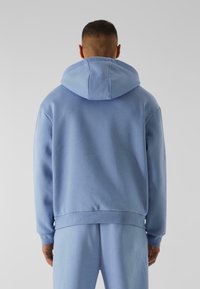 Lys blå hættebluse med en blød tekstur og ribbede manchetter. Har en stor hætte og minimalistisk design. Ingen synlige logoer eller mønstre.