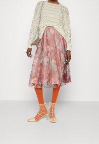 Jupe plissée rose clair à motif floral, associée à un pull en maille ivoire et des collants orange, ainsi que des bottines beiges semi-transparentes.