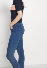 Jeans ajustados azules de denim, con una cintura a media altura, cinco bolsillos y una textura suave, con un sutil detalle de logo bordado.