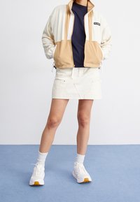 Krämen och beige zip-up jacka med sidofickor, ihop med en marinblå skjorta och en vit denimkjol, bärs med vita sneakers och ribbade strumpor.