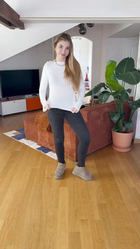 Haut à manches longues blanc, jean skinny noir, bottines grises à la cheville. Parquet en bois, plante d'intérieur et canape marron dans une pièce bien éclairée.