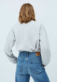 Felpa grigia chiara con maniche lunghe abbinata a jeans in denim blu, con una toppa in pelle marrone etichettata "PEPE JEANS" sulla cintura posteriore.