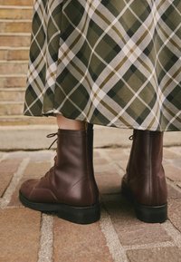 Botine de luptă din piele maro, cu șireturi, cu un finisaj mat, tălpi din cauciuc negru și cusături contrastante, asociate cu o fustă cu model în carouri.