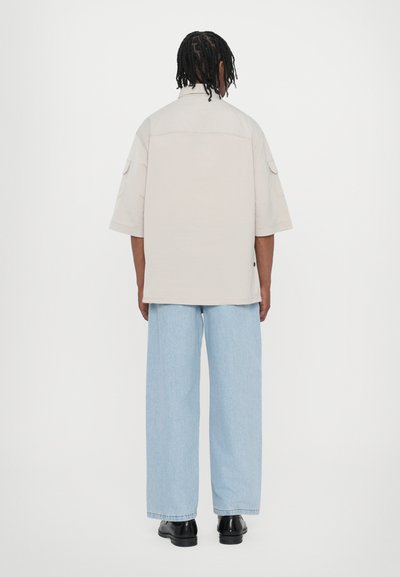 Chemise beige à manches courtes avec une coupe décontractée, poches latérales et pantalon en denim large bleu clair. Des chaussures noires complètent l'ensemble.