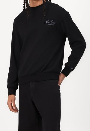 Mężczyzna ubrany w czarną bluzę z długim rękawem z napisem "New Era Since 1920" na piersi oraz czarne spodnie, stojący z jedną ręką w kieszeni.