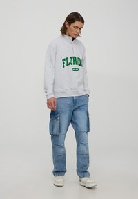 Felpa grigia con testo "FLORIDA" verde e colletto alto, abbinata a jeans cargo azzurri chiari con molteplici tasche.