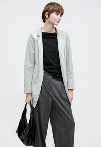 Világosszürke blézer gallérral, fekete felsővel és bőszárú szürke nadrággal párosítva. Fekete táskát tart a jobb kézben.