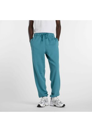 Mann trägt türkisfarbene Jogginghose mit Kordelzug und weiße Sneaker, Hände in den Taschen, vor einem einfarbigen Hintergrund.