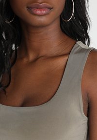 Närbild av en person som bär stora hoopörhängen och en ärmlös grå topp, som visar nacke, axlar och övre bröst.