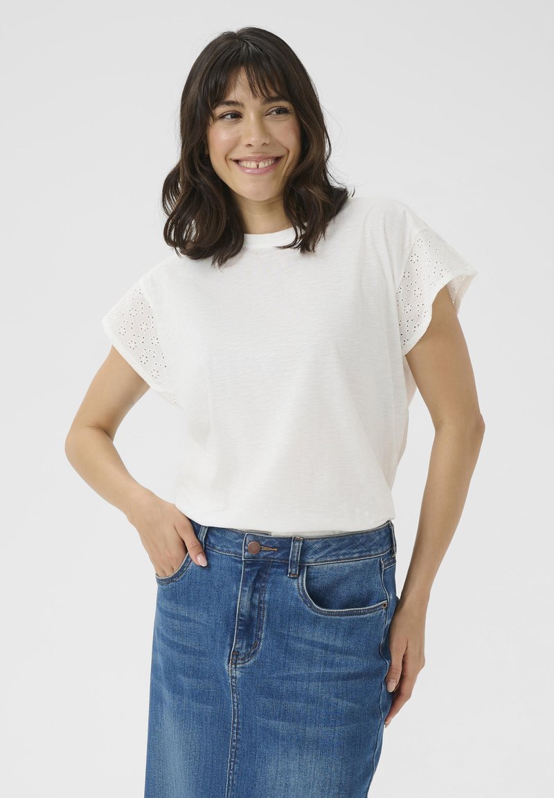 Haut blanc à manches courtes avec des accents en dentelle ajourée, associé à une jupe en denim bleu, mettant en valeur le design décontracté et texturé.