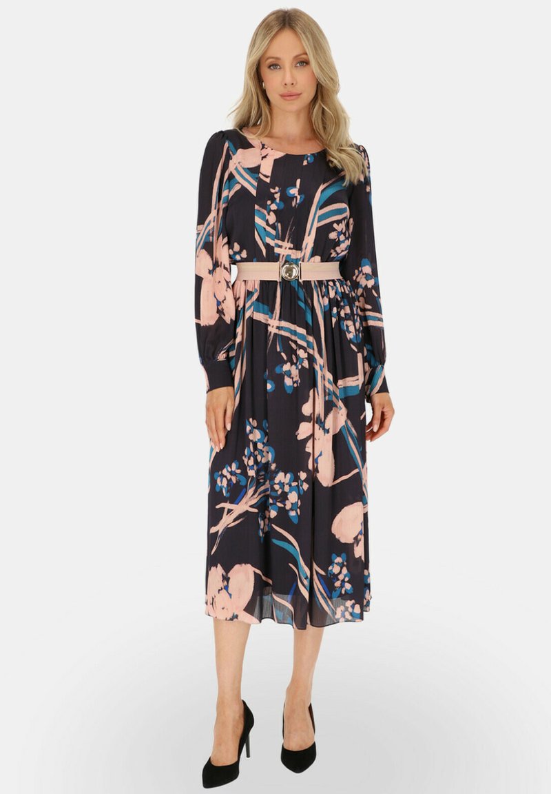 Długa rękawowa, granatowa sukienka w kwiaty z różowym i niebieskim wzorem, z zaznaczoną talią, dopasowanym paskiem i plisowanym dołem. Czarne szpilki dopełniają stylizację.