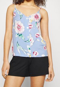 Lichtblauwe cami-top met bloemenprint, met spaghettibandjes en knoopsluiting, versierd met roze en groene bloemen. Gecombineerd met zwarte broek met pinstripes.