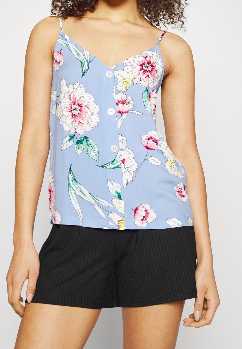 Lichtblauwe cami-top met bloemenprint, met spaghettibandjes en knoopsluiting, versierd met roze en groene bloemen. Gecombineerd met zwarte broek met pinstripes.