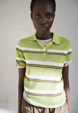 Joven de piel oscura con una camiseta tipo polo de rayas verdes y blancas, de pie contra una pared lisa y clara.