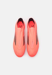 Jasnoróżowe korki do piłki nożnej typu slip-on z teksturowaną powierzchnią, z czarnymi akcentami i aerodynamicznym kształtem. Brak widocznych sznurowadeł.