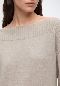 Femme portant un pull en maille côtelée beige décolleté aux épaules, montrant la clavicule et la moitié inférieure du visage avec une couleur naturelle des lèvres.