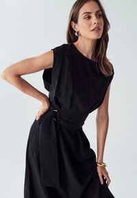 Črna obleka iz teksturiranega blaga, s širokim izrezom in zavezanim pasom s krožno sponko. Gladek, tekoč silhuet.