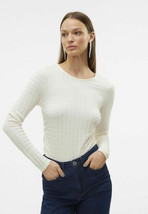 Hvit langermet topp med et teksturert mønster og rund hals, kombinert med mørkeblå denimjeans. Stoffet ser mykt og tettsittende ut.
