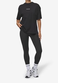 Person wearing black oversized t-shirt, black leggings, white socks with text, and white New Balance sneakers standing on white background.