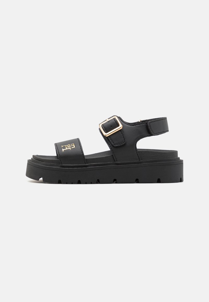 Svart sandal laget av syntetisk materiale med to justerbare stropper, en kraftig såle og en gullfarget spenne. Horisontale riller på sålen.