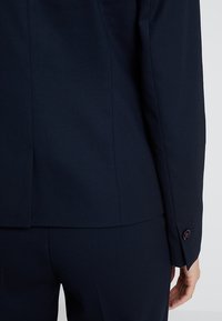Mörkblå blazer med texturerat tyg, strukturerad design och detaljer med enstaka knapp vid ärmen. Skräddarsydd passform med en liten slits bak.