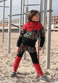 Kolorowy dres z czerwonymi, czarnymi i kwiatowymi wzorami. Bluza z kapturem ma czerwoną górną część, natomiast spodnie mają kwiatowy i czarny wzór.