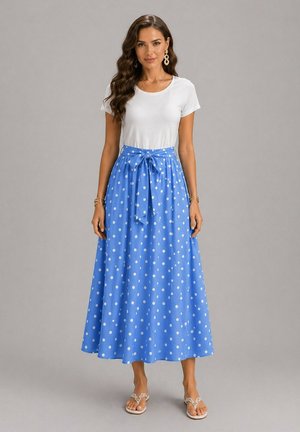 Femme aux longs cheveux ondulés portant un haut blanc à manches courtes, une jupe midi bleue à pois taille haute avec un nœud, et des sandales blanches, debout sur un fond gris.