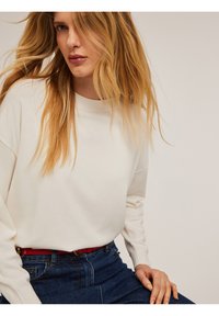 Suéter crema de manga larga, con cuello redondo y puños acanalados, combinado con unos pantalones vaqueros de talle alto en azul y un cinturón rojo.