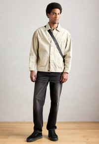 Bărbat cu păr creț, purtând o jachetă bej, un tricou negru, blugi negri, adidași negri și o geantă crossbody neagră, stând pe un pardoseală din lemn.