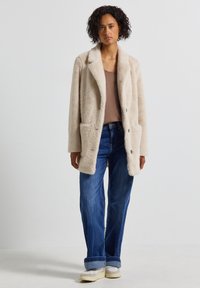 Manteau en fausse fourrure beige avec un col à revers, deux poches avant et une coupe décontractée, associé à un jean bleu décontracté et des baskets blanches.