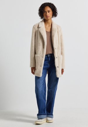 Beige nepbontjas met een omgeslagen kraag, twee voorzakken en een ontspannen pasvorm, gecombineerd met casual blauwe jeans en witte sneakers.