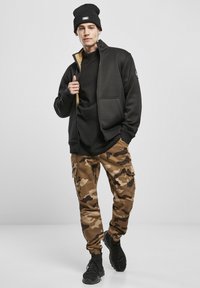 Juodas zip-up švarkas su chaki pamušalu, derinamas su juodu marškinėliu ir rudais kamufliažo krovinių kelnėmis. Juodi sportiniai bateliai ir juodas kepurėles užbaigia įvaizdį.