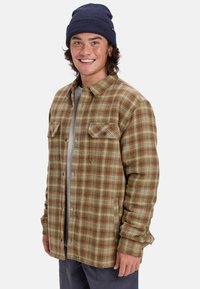 Braunes und beiges kariertes Flanellhemd mit Brusttaschen, kombiniert mit einem grauen T-Shirt. Das Model trägt eine marineblaue Mütze und zeigt einen lässigen Stil.