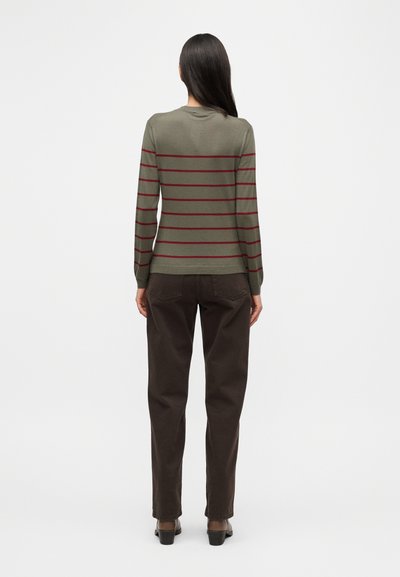 Πράσινο πουλόβερ με μακριά μανίκια και κόκκινες οριζόντιες ρίγες, με στρογγυλή λαιμόκοψη και ριμπ μανσέτες, συνδυασμένο με καφέ παντελόνι και παπούτσια με τακούνι.