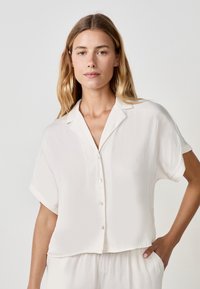 Blusa bianca a maniche corte con colletto e chiusura a bottoni, realizzata in un tessuto leggero e morbido, con una vestibilità rilassata.