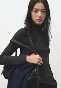 Black ribbed turtleneck top, paired with a black bag and a dark blue sweater. Model has long dark hair and is standing against a white wall.
