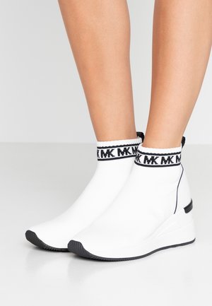 Baskets chaussettes blanches à talon compensé avec semelles noires et logo "MK" en bordure, portées sur des jambes nues sur un fond gris uni.