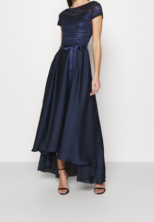 Frau trägt ein dunkelblaues, strukturiertes Kurzarmtop mit passendem Satin-High-Low-Rock und schwarzen Absatzschuhen mit Knöchelriemen.