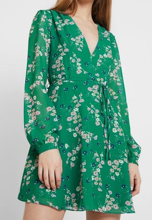 Frau trägt ein grünes Blumenkleid mit langen Ärmeln, tiefem V-Ausschnitt und Taillenbindegürtel, stehend vor einem schlichten Hintergrund.