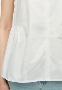 Λευκή αμάνικη μπλούζα με λεπτομέρεια σούρας στη μέση, συνδυασμένη με ανοιχτόχρωμο τζιν παντελόνι, μερική θέα του αριστερού βραχίονα.